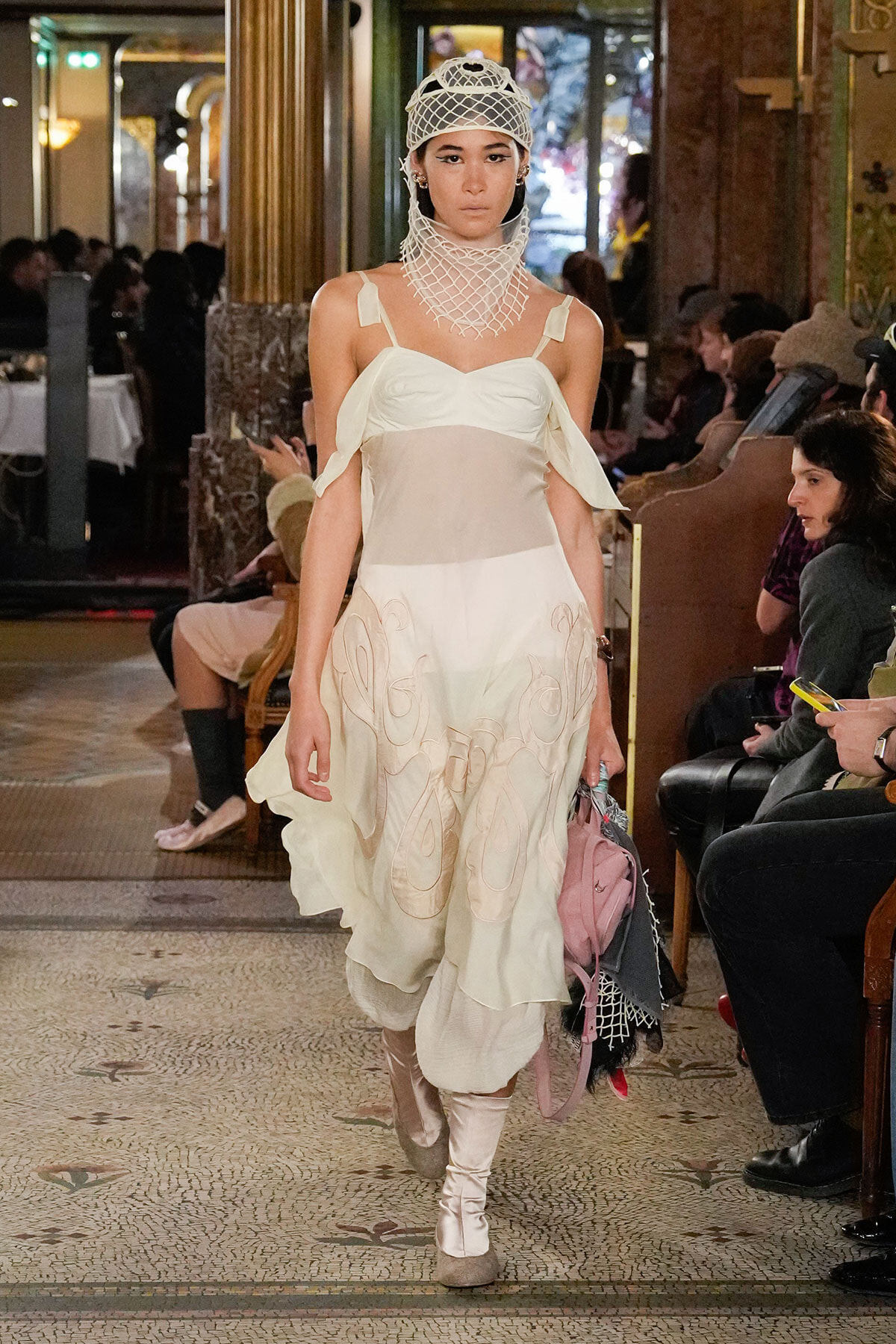 Model walks a runway in an ivory layered dress with shoulder straps and a sculptural mesh neck/hood, indoors at a fashion show.