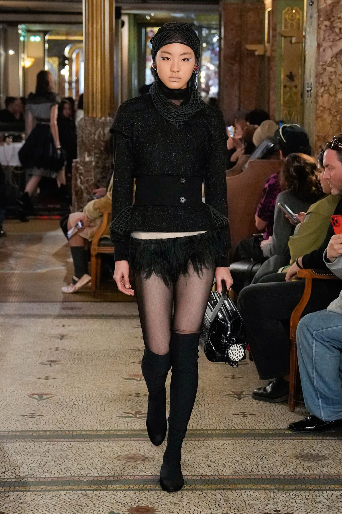 Model walks a runway in a black textured outfit with a wide belt, feathered hem, sheer tights and thigh-high boots inside a grand venue.