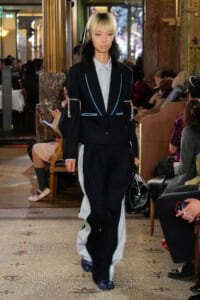 Model walking the runway in a dark pinstripe suit with blue piping, layered over a light striped shirt, indoors at a fashion show venue.