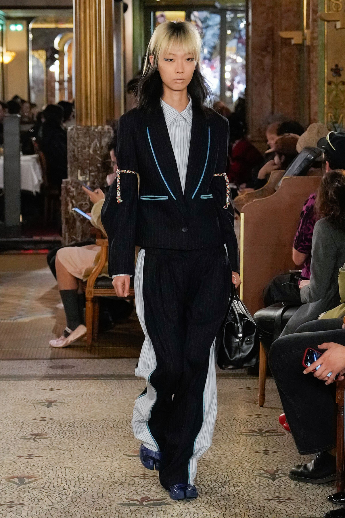 Model walking the runway in a dark pinstripe suit with blue piping, layered over a light striped shirt, indoors at a fashion show venue.