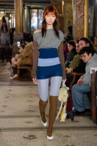 Model walks a runway in a gray off-shoulder top, blue color-block leggings, and brown thigh-high boots as spectators watch in a ornate venue.