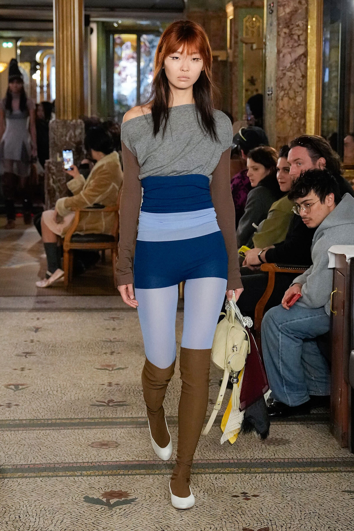 Model walks a runway in a gray off-shoulder top, blue color-block leggings, and brown thigh-high boots as spectators watch in a ornate venue.