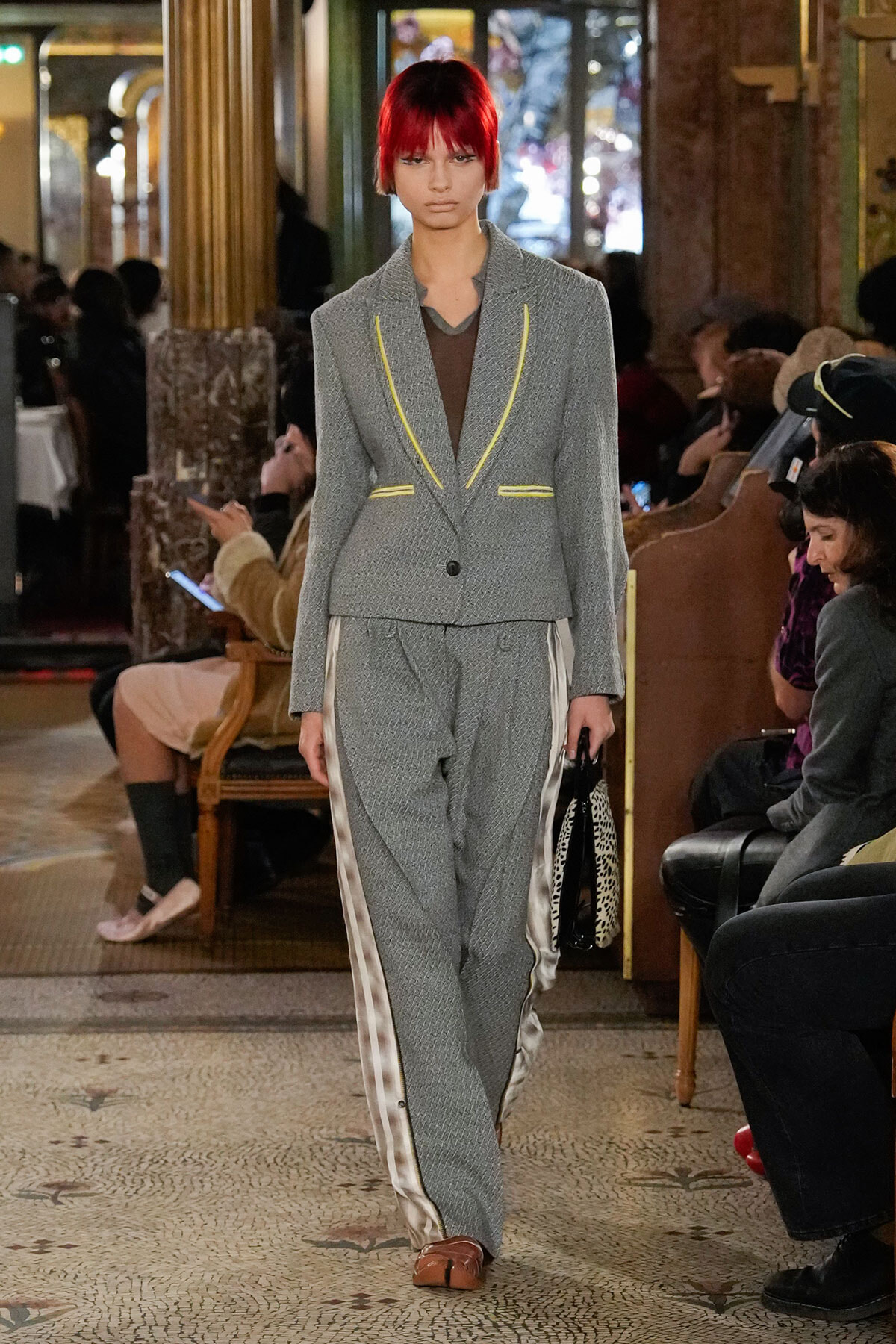 Model walking a fashion runway in a gray suit with yellow piping and satin side stripes, in a ornate indoor venue with audience watching.