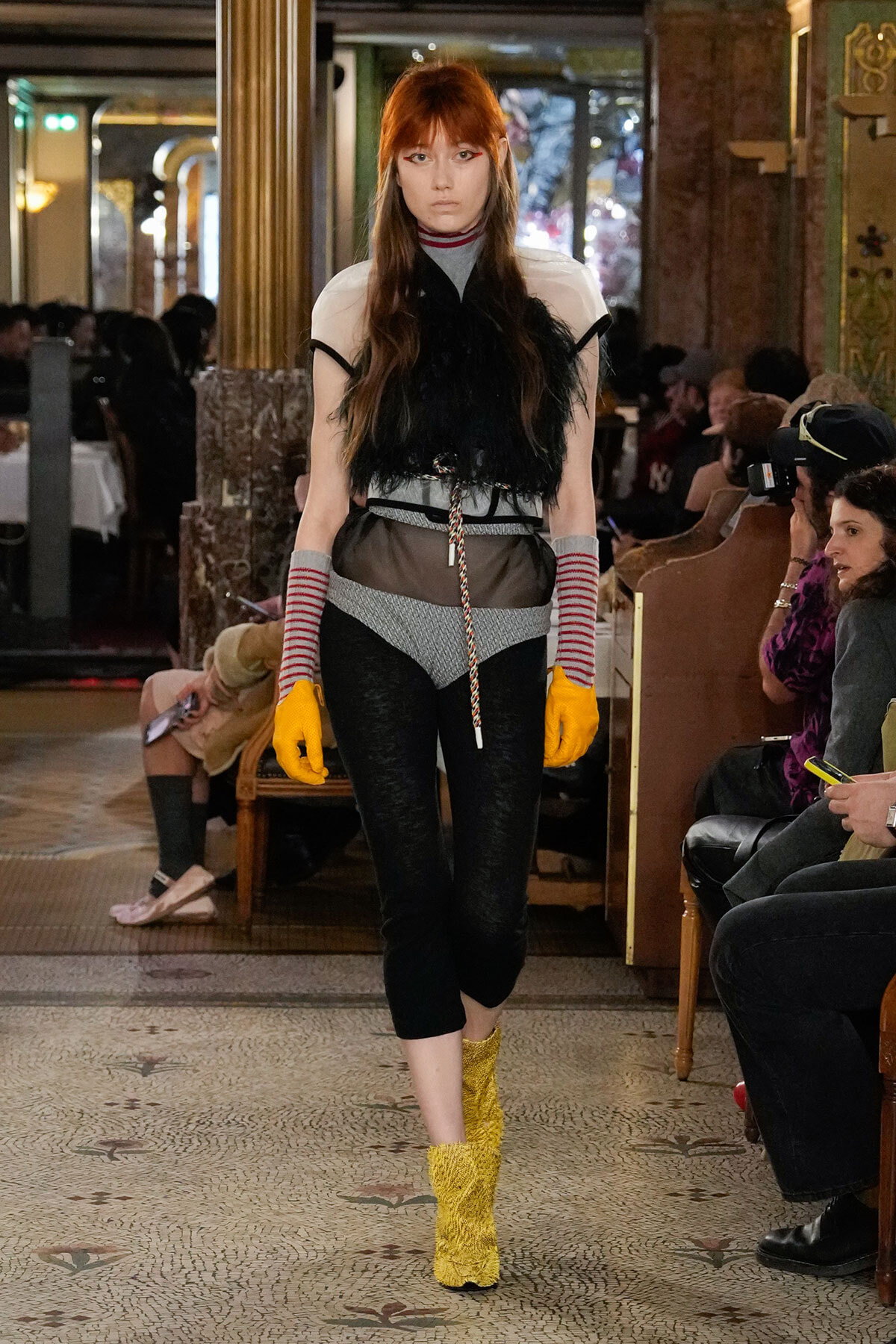 Model walks the runway in a layered outfit: black feathered top over gray turtleneck, striped arm warmers, sheer waist panel, and bright yellow knit boots.