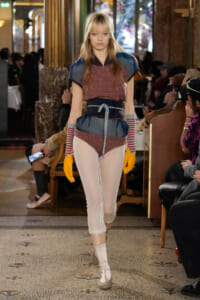Model walks a runway wearing a layered maroon plaid top with blue shoulder panels, a blue belt, and a rope sash over sheer white tights and beige ankle boots, with bright orange gloves and striped arm warmers.