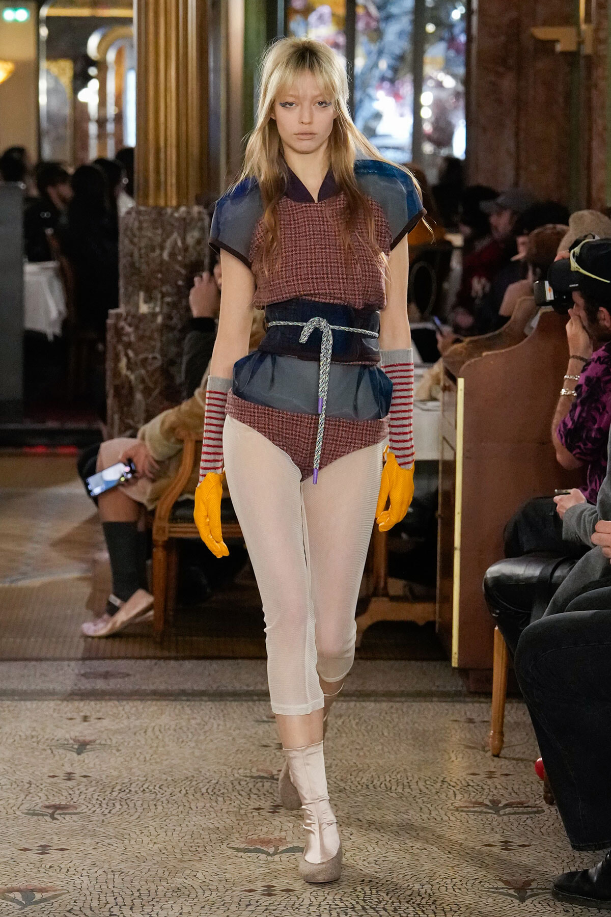 Model walks a runway wearing a layered maroon plaid top with blue shoulder panels, a blue belt, and a rope sash over sheer white tights and beige ankle boots, with bright orange gloves and striped arm warmers.