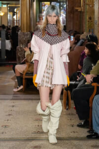 Runway model walking center stage in a pale pink oversized shirt with a black fishnet yoke, striped sleeves, a plaid dress, bright yellow gloves, and tall off-white boots.