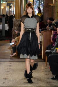 Model walking a runway in a gray knit top with sheer black sleeves and a voluminous black bubble skirt, cinched with a rope belt in a busy venue.