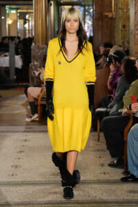 Model walking a fashion runway in a bright yellow V-neck dress with black trim, paired with black gloves and slouchy boots, inside an ornate venue with seated audience