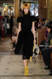 Model walks the runway in a black puff-sleeve top and feathered black skirt, with yellow-trim details and bright yellow knit boots.