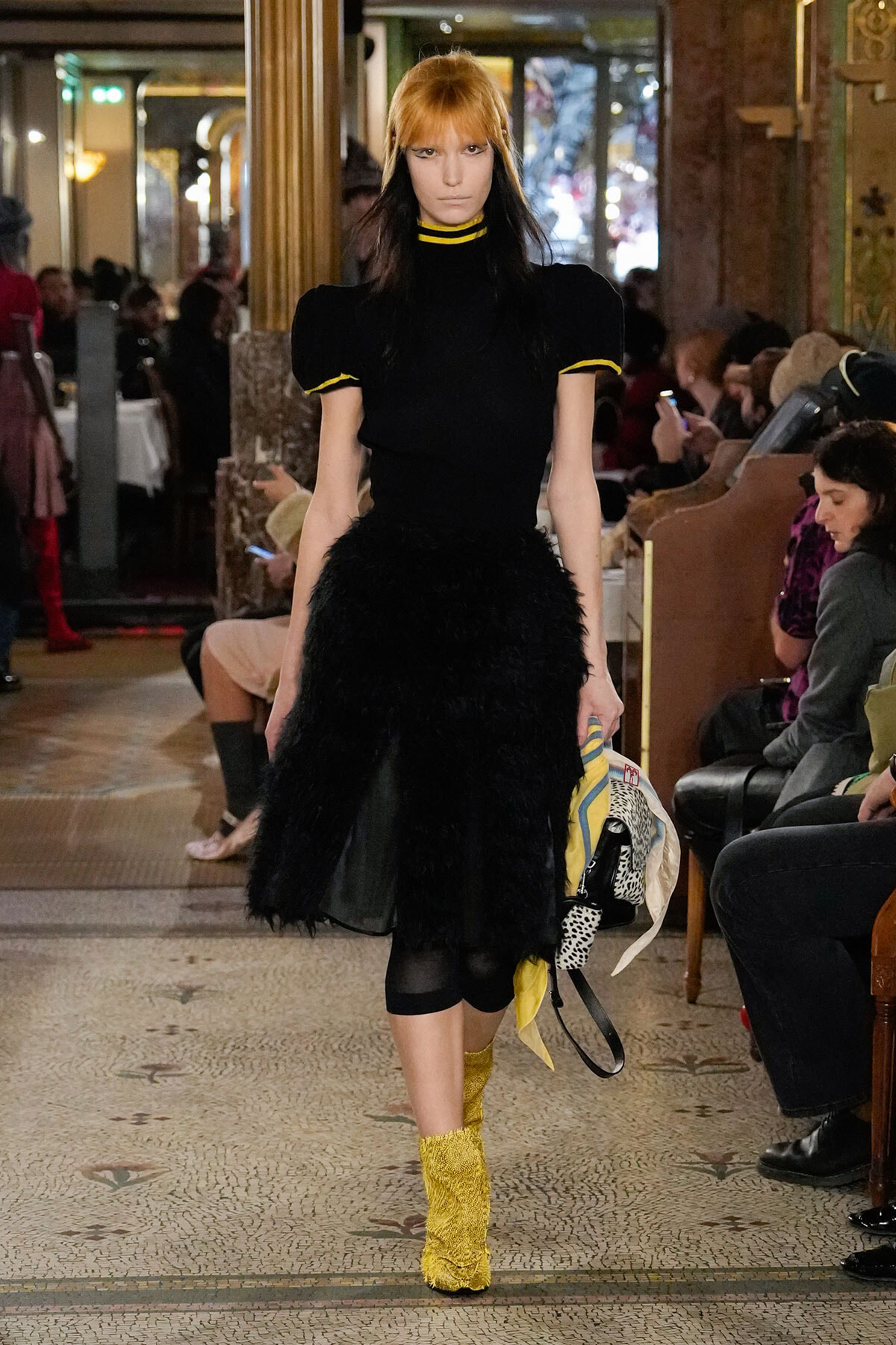 Model walks the runway in a black puff-sleeve top and feathered black skirt, with yellow-trim details and bright yellow knit boots.