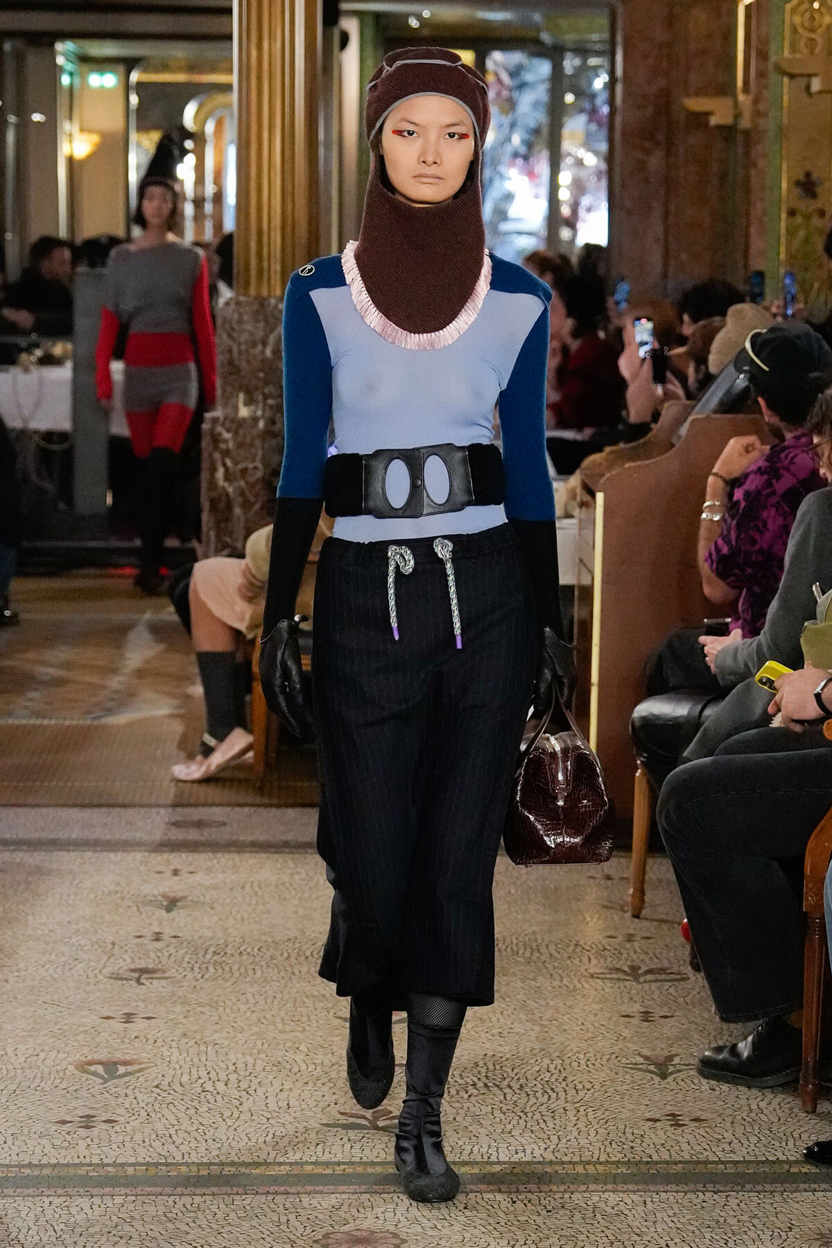 Model walking a runway in a blue and light-blue color-block top, brown knit hood and scarf, black belt with oval buckle, pinstripe trousers, gloves, boots, and a burgundy handbag; audience in background