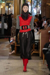 Model walking runway in a red sleeveless top over gray sleeves, black textured pants, leopard belt, red boots and black headwear in a fashion show venue.