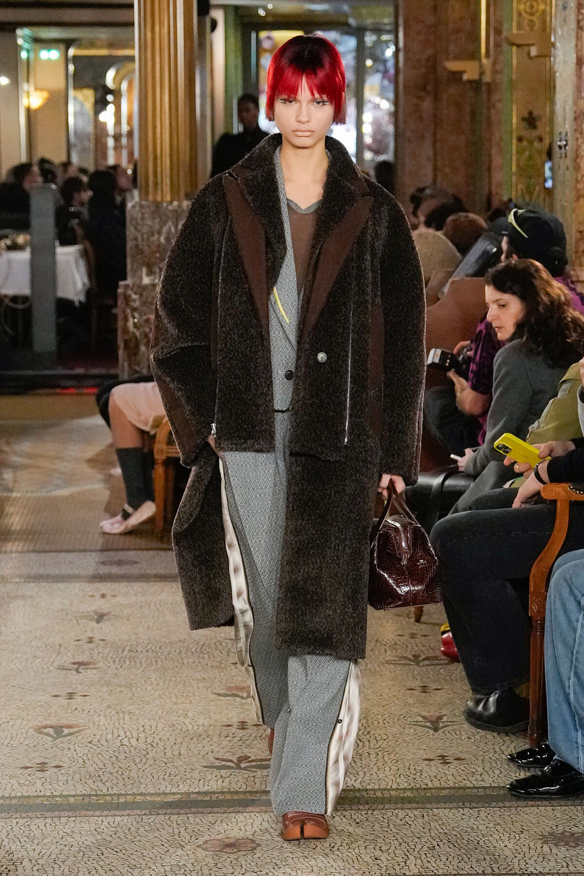 Model walking a fashion show in a plush brown coat over a grey suit, red-and-black hair, carrying a maroon handbag in a luxurious venue.