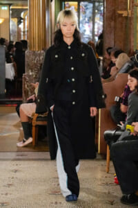 Model walking the runway in a long black coat with large pockets and silver buttons, inside a busy venue with seated spectators.