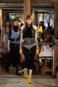Model walks in a fashion show wearing a fur top, striped sleeves, and bright yellow gloves with fuzzy yellow boots in a cafe-like venue.