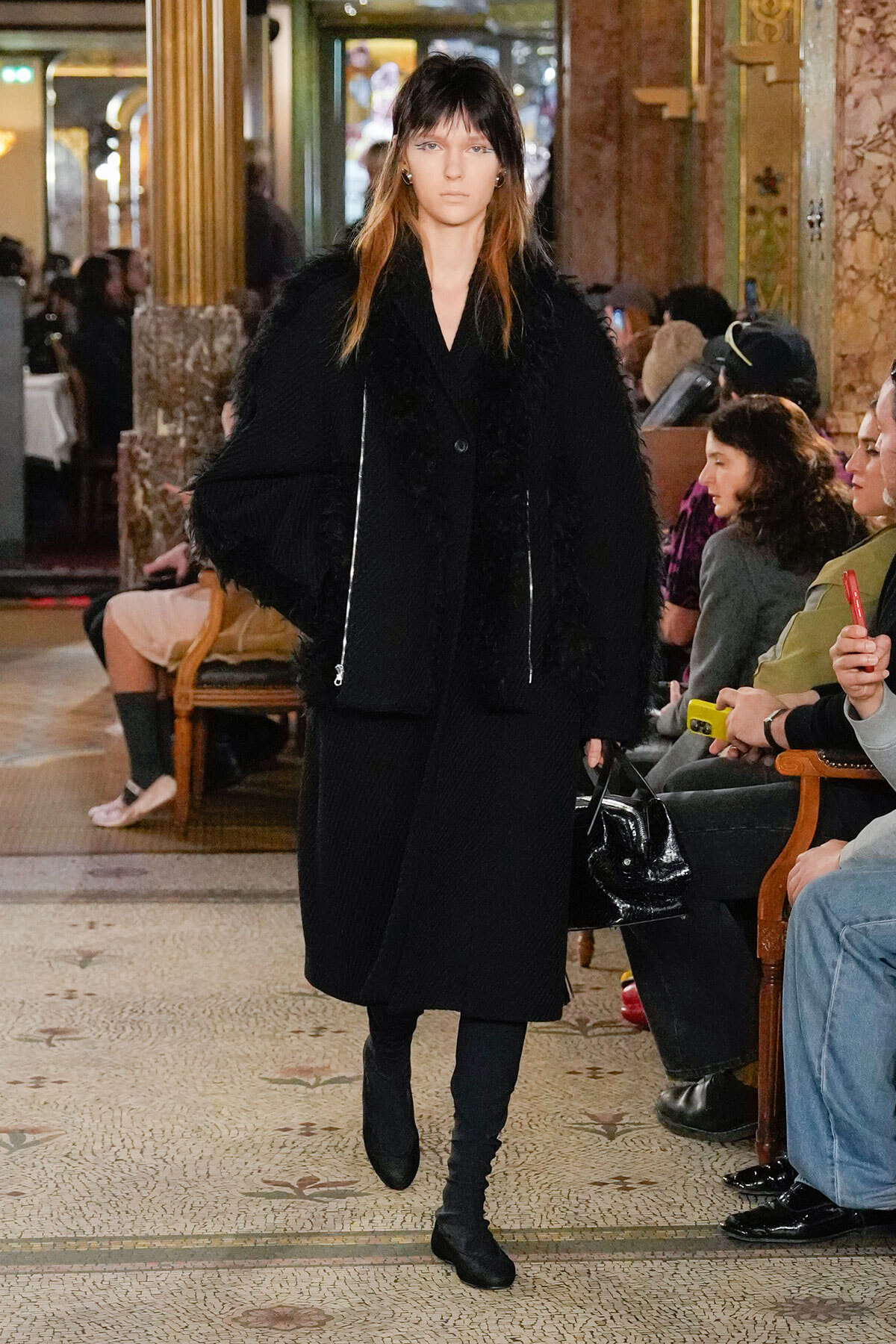Model walking a runway in a long, black textured coat with a fluffy front, inside an ornate lobby with seated spectators nearby.