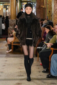 Runway model wearing a dark textured coat, sheer tights, and knee-high boots at a fashion show.