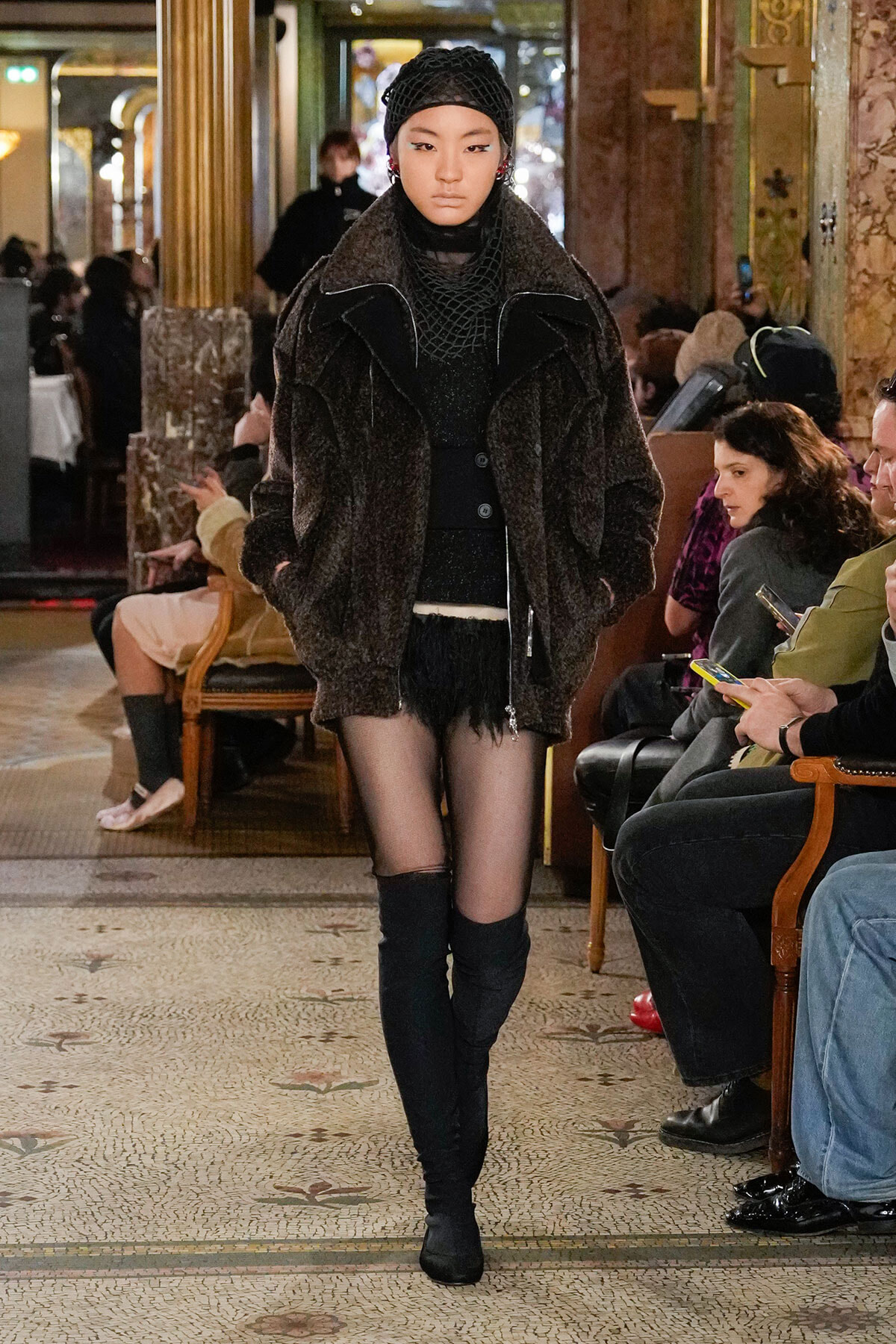 Runway model wearing a dark textured coat, sheer tights, and knee-high boots at a fashion show.
