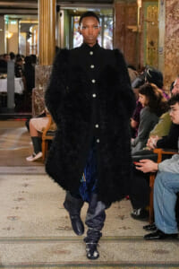 Model walks the runway in a voluminous black fuzzy coat with silver button row, blue patterned pants, and black boots in a grand venue.