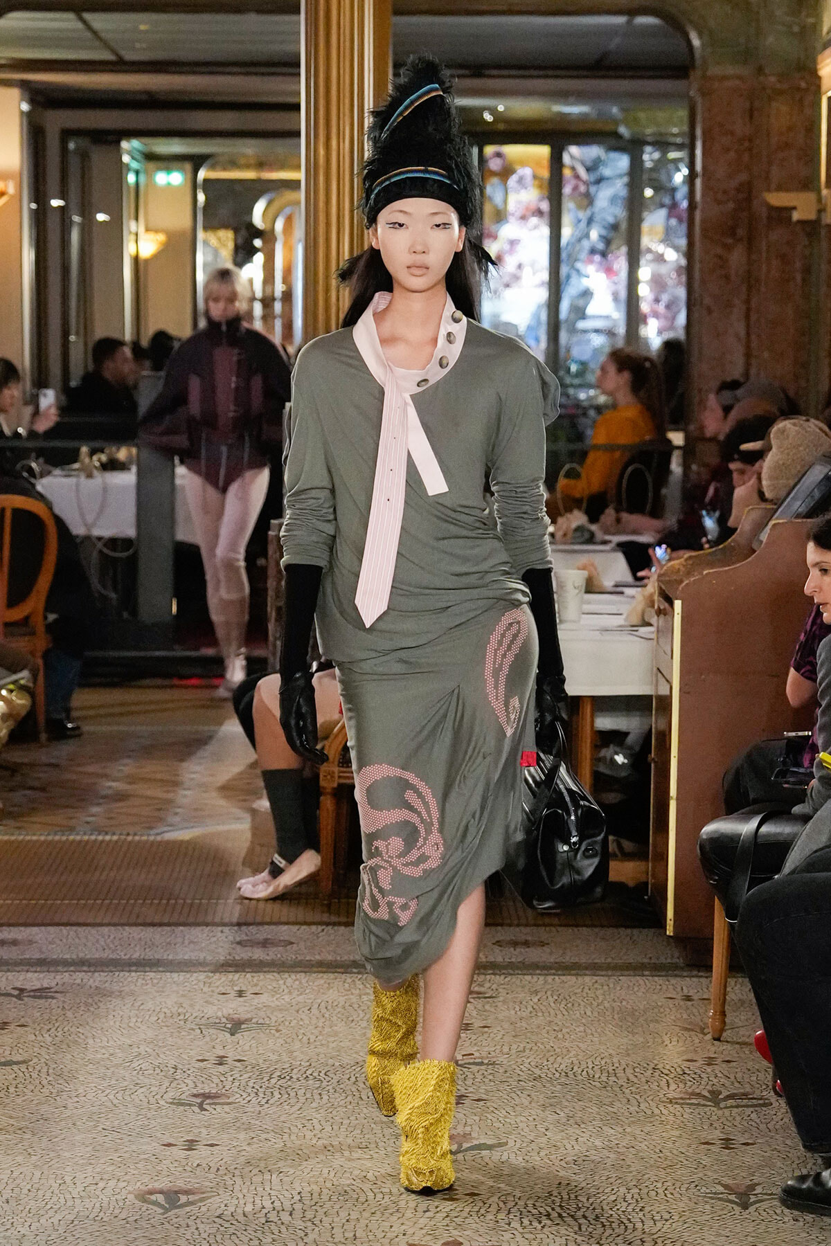 Model walking a couture runway in a loose olive-green dress with pink scarf, black gloves, and yellow fuzzy boots in a lavish hotel venue.