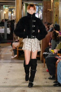 Model walks runway in a black fur coat with silver toggles, paired with striped beige shorts and tall black boots.