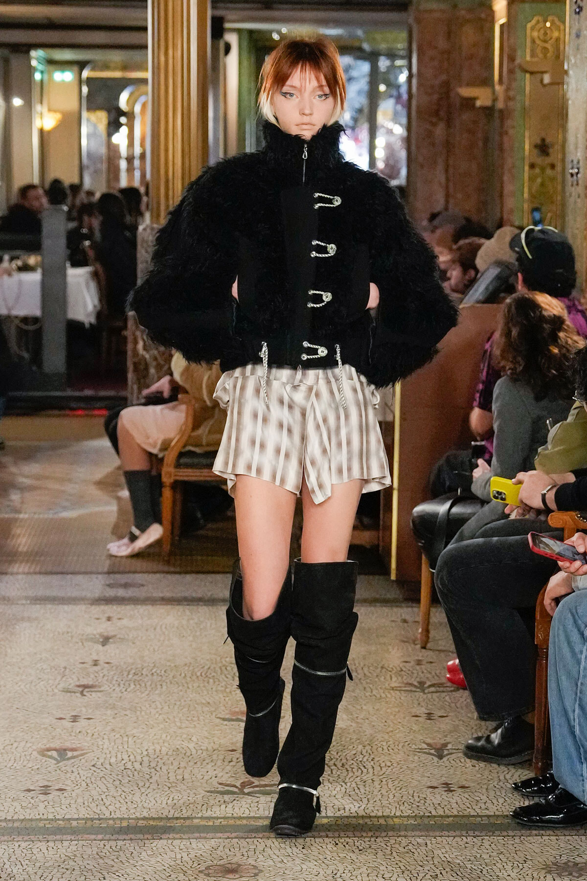 Model walks runway in a black fur coat with silver toggles, paired with striped beige shorts and tall black boots.