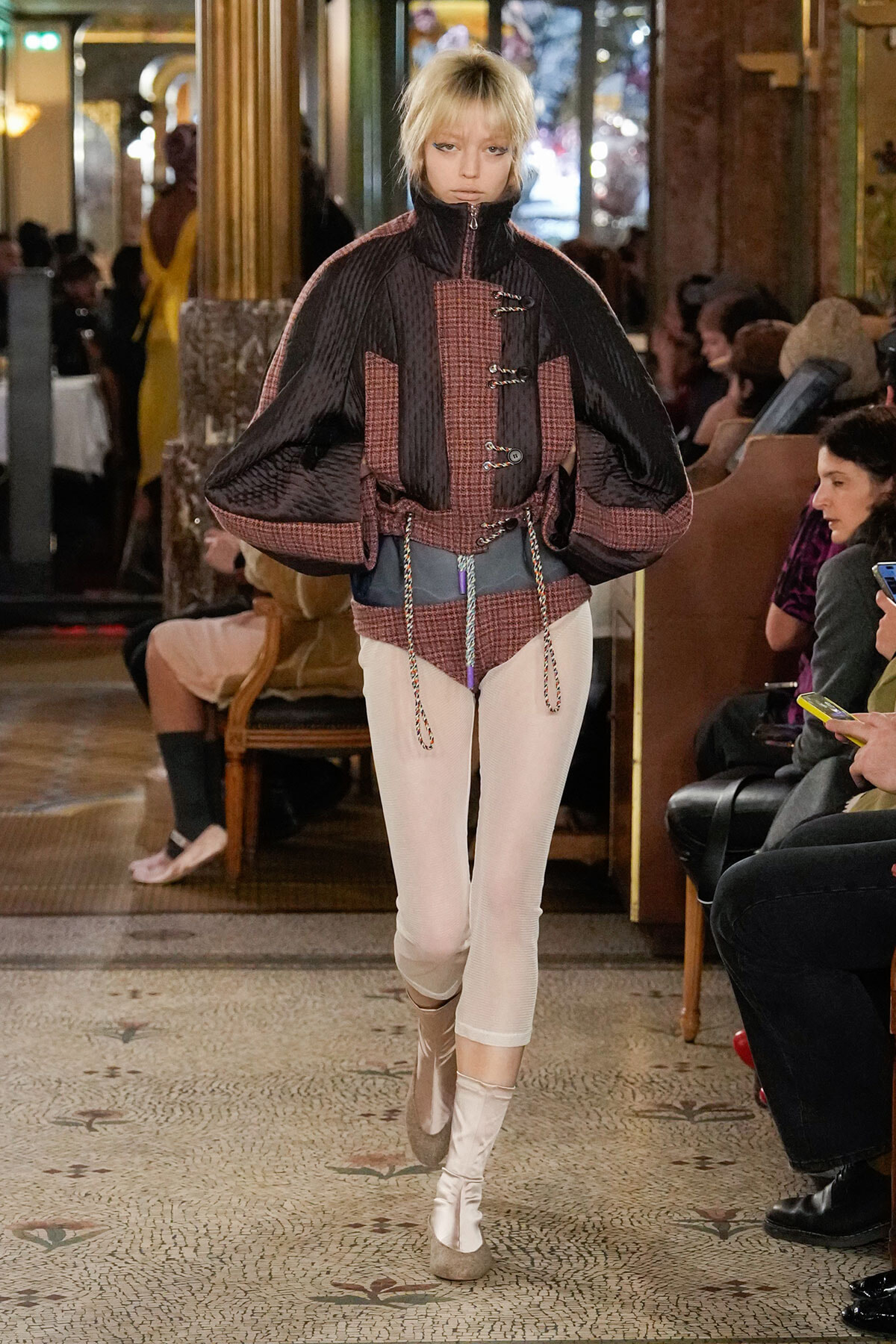 Model walking a fashion show runway in an oversized burgundy plaid jacket with dramatic puff sleeves, white tights, and light boots; audience seated along the sides.