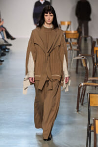 Model on a runway wearing a tan layered coat with a belt over a striped shirt and wide-legged pants, neutral tones.
