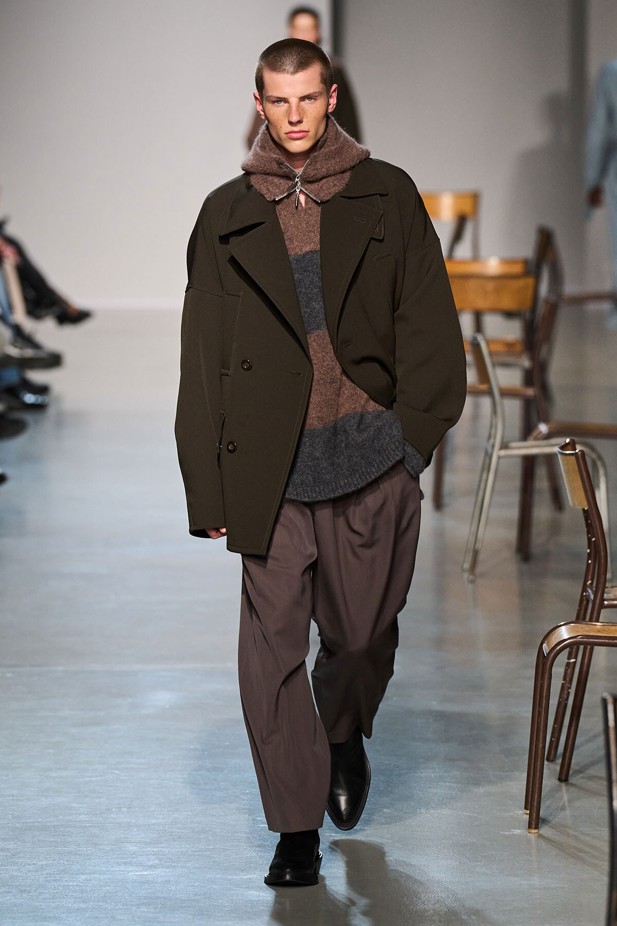 Male model walking the runway in an oversized dark olive coat layered over a brown-gray knit sweater and loose brown trousers, black boots visible.