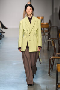 Model walking a runway in a pale lime double-breasted blazer over a dark brown shirt and wide brown trousers on a minimalist catwalk.