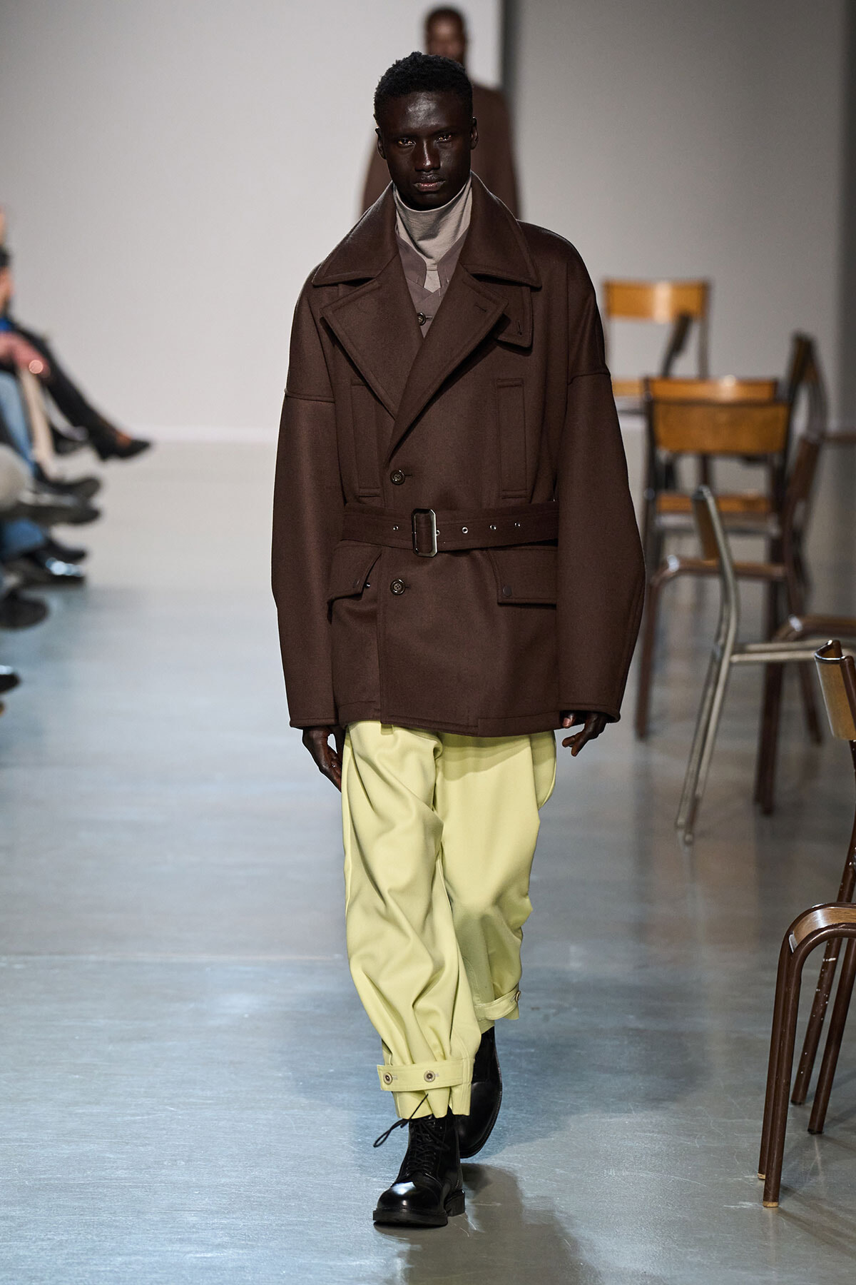 Runway look: model wearing a brown belted oversized coat, beige turtleneck, pale yellow pants, and black boots.