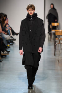 Male model walks a runway in an all-black, layered outfit: double-breasted jacket, oversized scarf, black pants, and boots, with audience along the catwalk