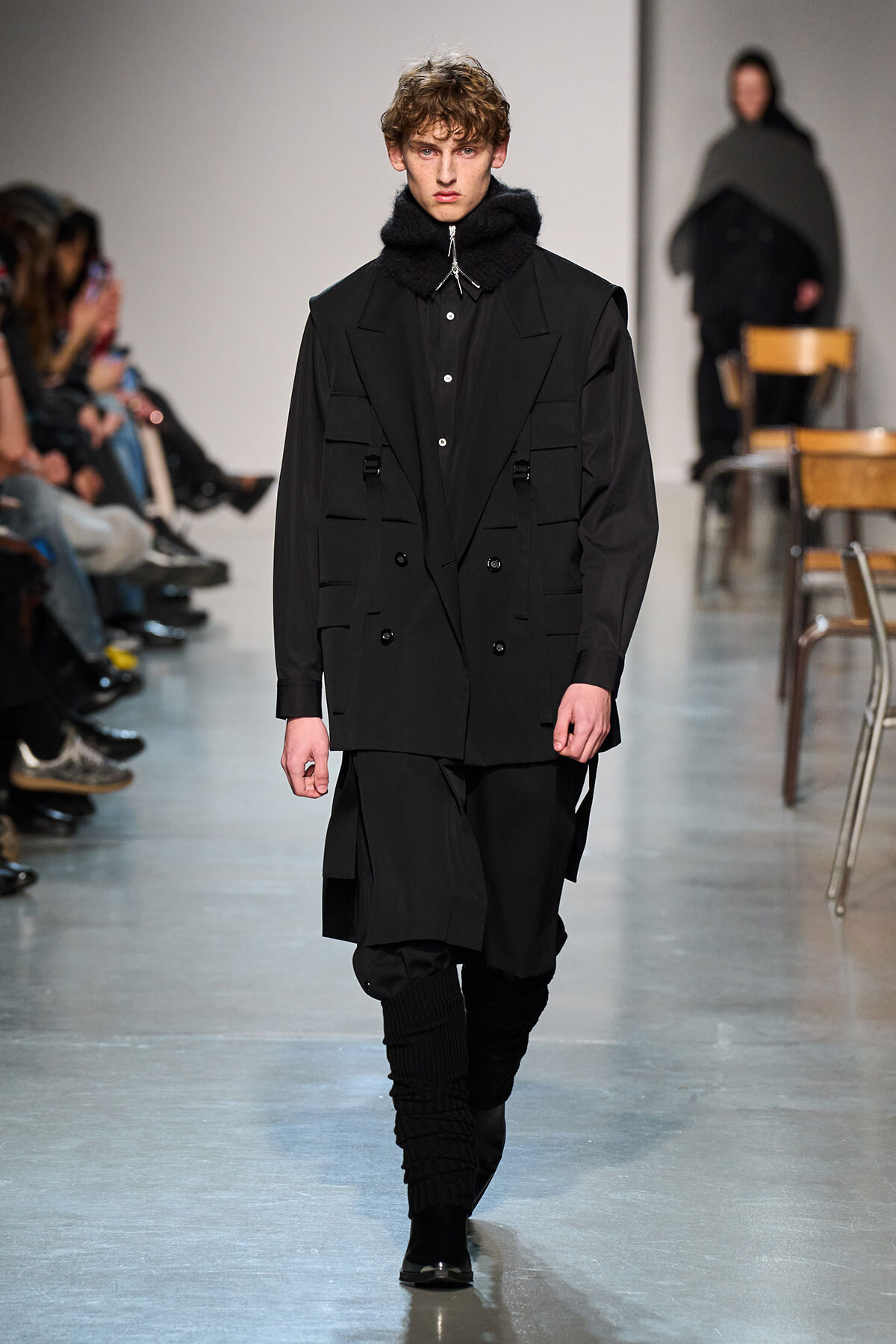 Male model walks a runway in an all-black, layered outfit: double-breasted jacket, oversized scarf, black pants, and boots, with audience along the catwalk