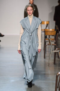 Model on a runway wearing a light blue pinstripe sleeveless belted coat over a beige turtleneck.