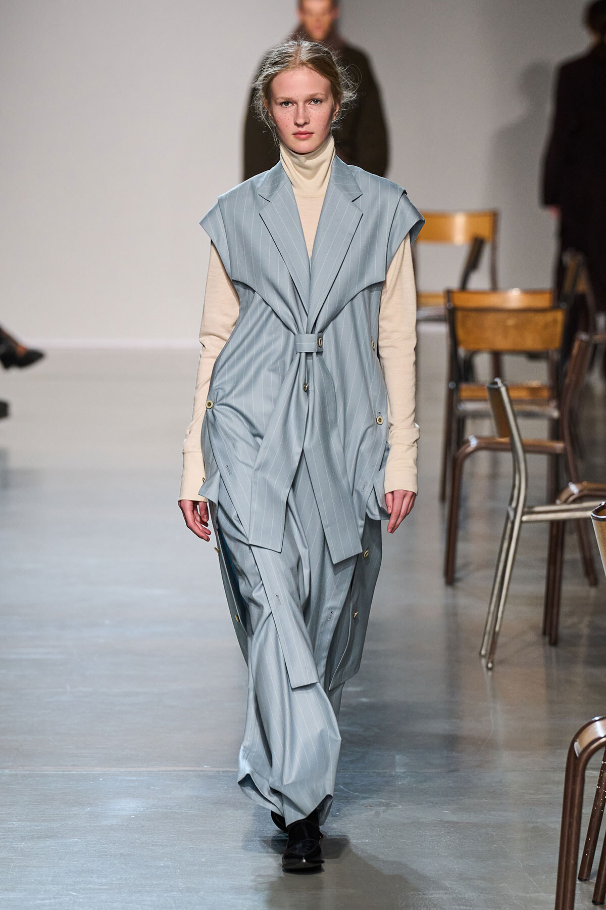 Model on a runway wearing a light blue pinstripe sleeveless belted coat over a beige turtleneck.