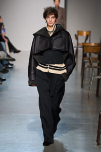 Model walks a fashion runway in a dark, layered coat with shearling accents over a cream striped sweater and wide black pants.