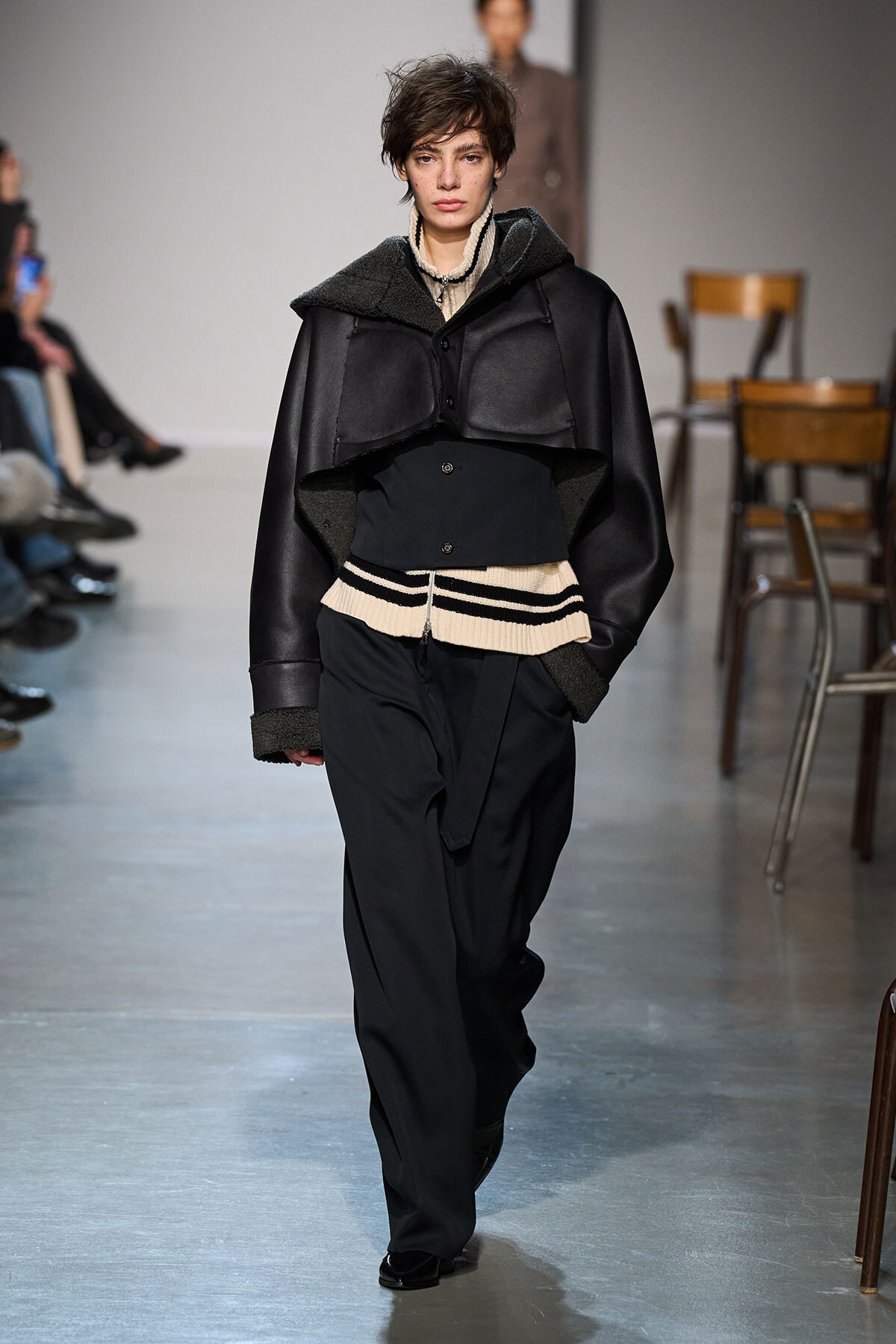 Model walks a fashion runway in a dark, layered coat with shearling accents over a cream striped sweater and wide black pants.