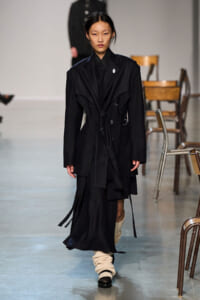 Model walks a fashion runway in a black, oversized pinstriped trench coat with a belt, layered panels, and long sleeves, paired with cream knit leg warmers and dark shoes.