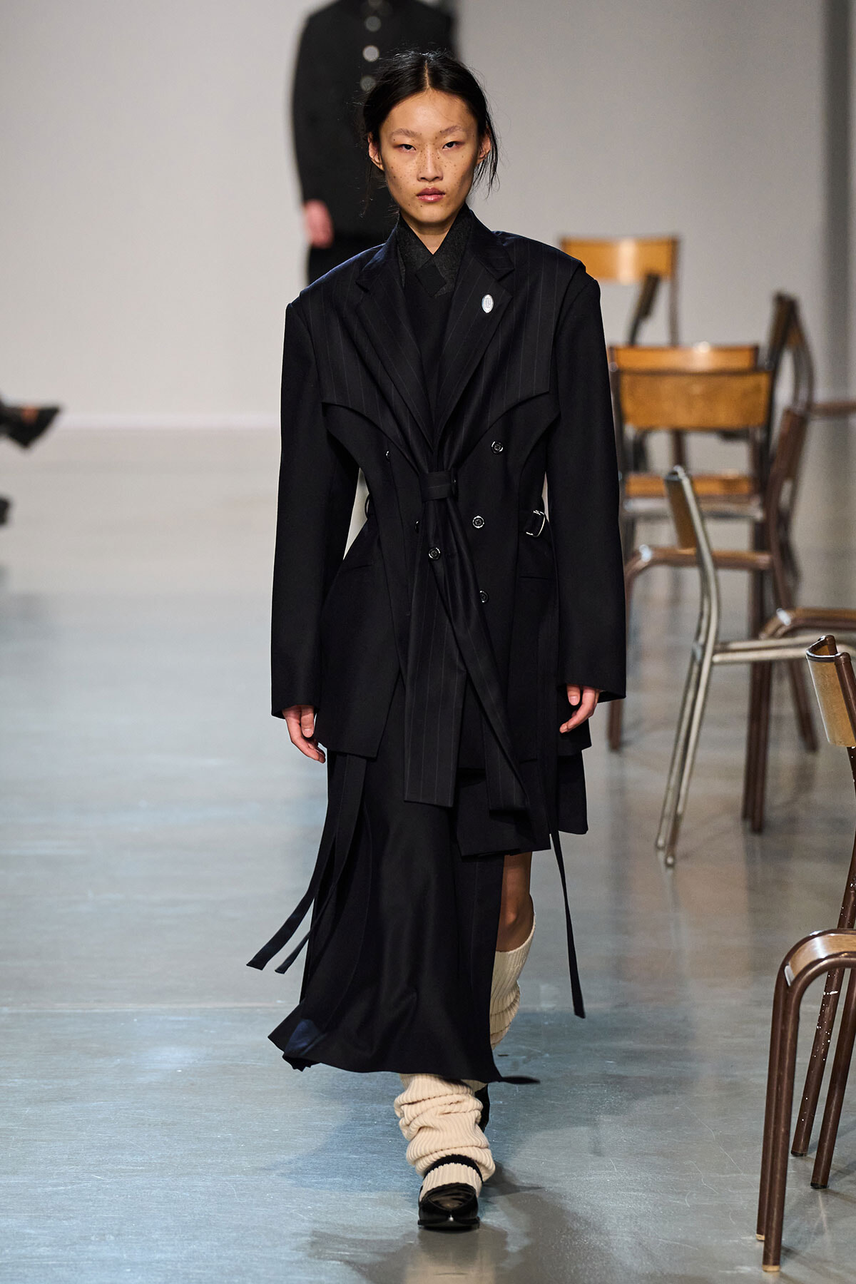 Model walks a fashion runway in a black, oversized pinstriped trench coat with a belt, layered panels, and long sleeves, paired with cream knit leg warmers and dark shoes.