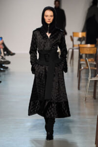 Model walks runway in a black quilted coat with hood, cinched waist belt, and tall boots.