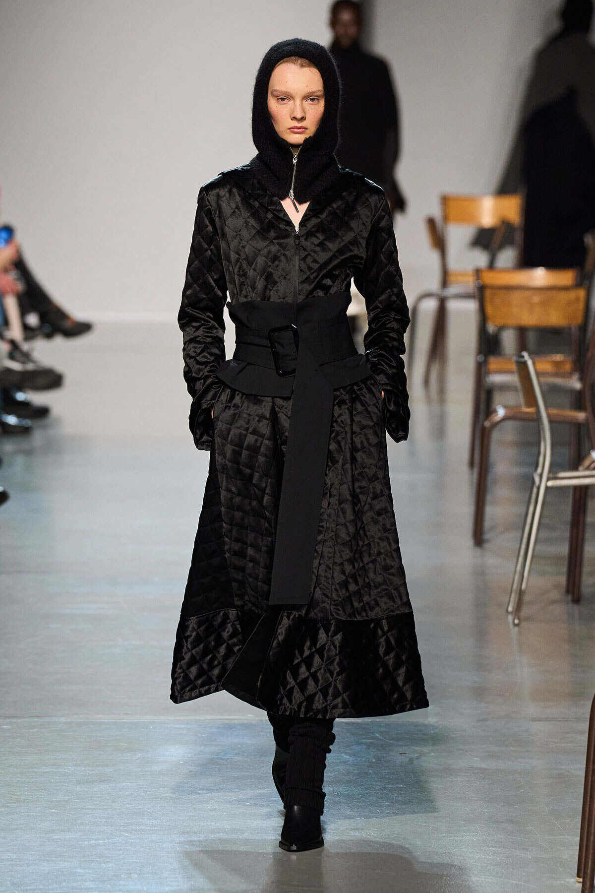 Model walks runway in a black quilted coat with hood, cinched waist belt, and tall boots.
