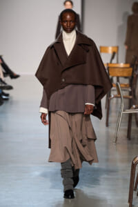 Model walks a fashion runway in a dark brown oversized cape over layered taupe outfits, with gray leg warmers and black shoes.