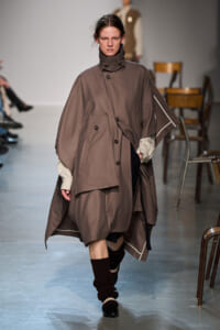 Model on a runway wearing an oversized taupe cape coat with a high collar and large front pockets, layered over striped cuffs. Showcasing a voluminous brown ensemble.