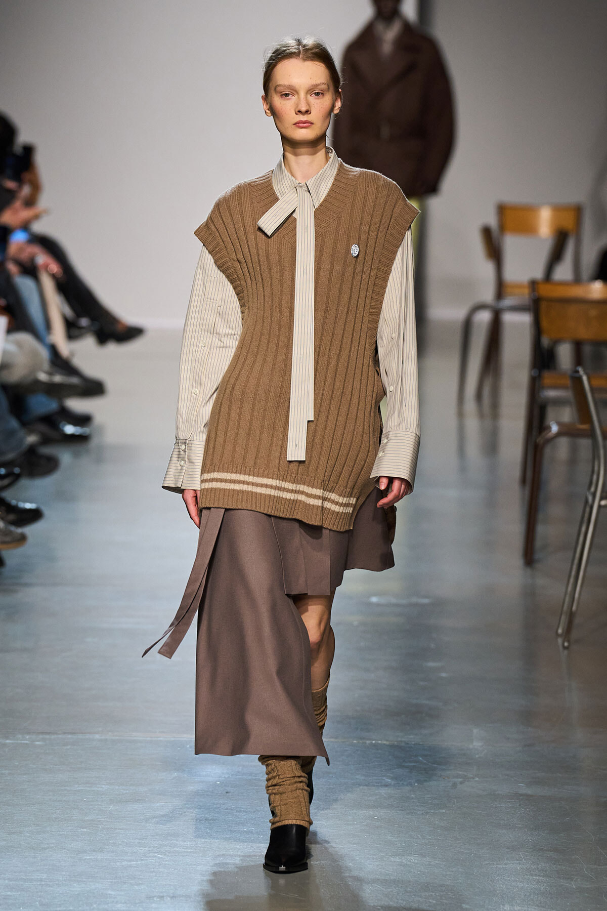 Model on a runway wearing a tan rib-knit sleeveless sweater layered over a beige striped shirt with a long tie, and a taupe skirt with leg warmers and ankle boots.