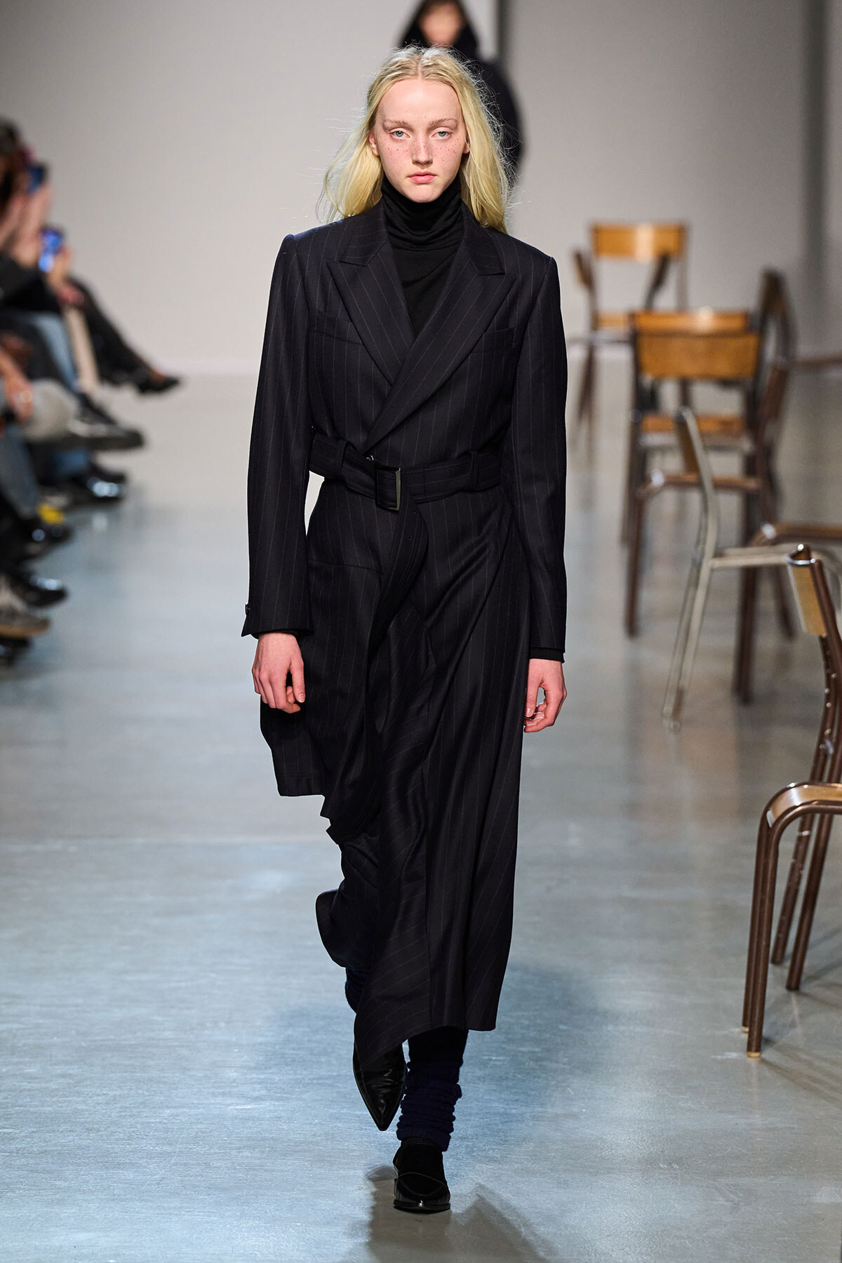 Female model walking a fashion show in a dark pinstripe suit with a belted blazer and flowing trousers on the runway