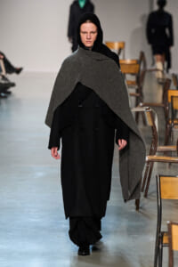 Model walks a fashion show wearing a long black coat with a large grey knit cape draped over her shoulders and hood. Chairs line the runway background.