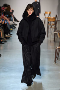 Model on a runway wearing an all-black look: hooded bomber jacket, turtleneck, and wide pinstripe trousers.