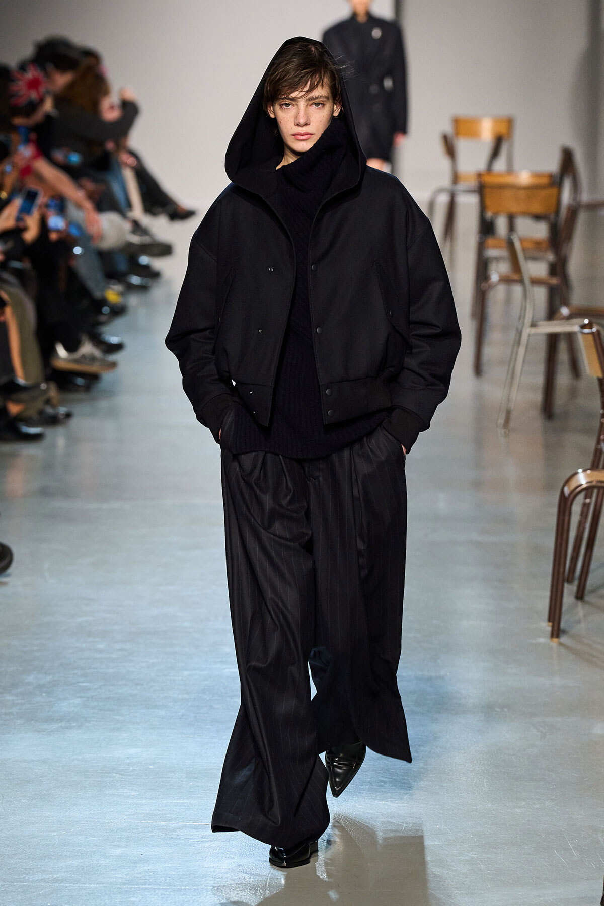 Model on a runway wearing an all-black look: hooded bomber jacket, turtleneck, and wide pinstripe trousers.
