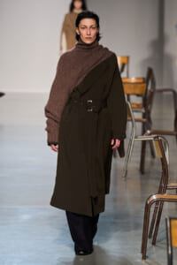 Model walking runway in a dark olive trench coat with a wide brown scarf draped over one shoulder.